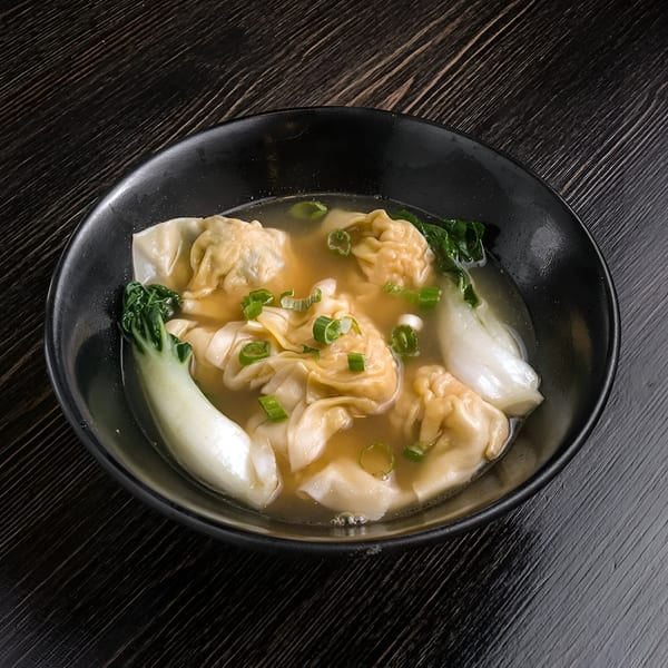 Chan Kee's wonton soup with bok choy and green onions in a black bowl on a dark wooden table.