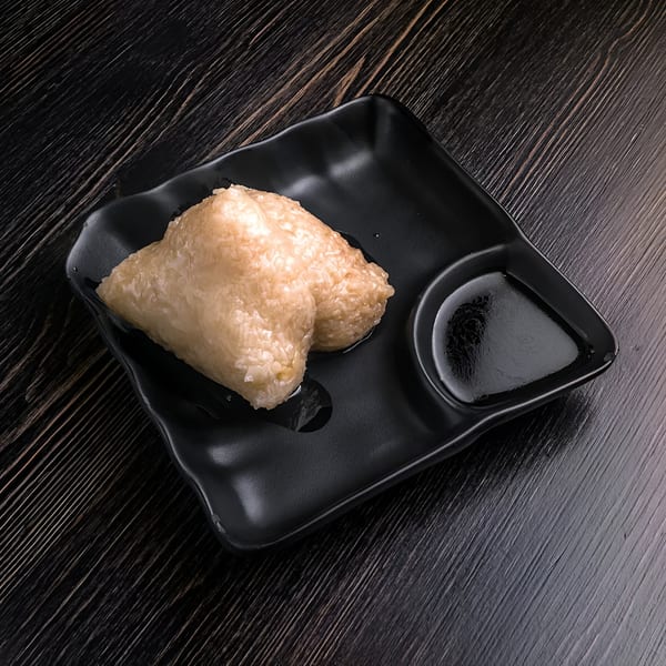 Two light brown zongzi, traditional Chinese sticky rice dumplings, on a black plate with soy sauce.